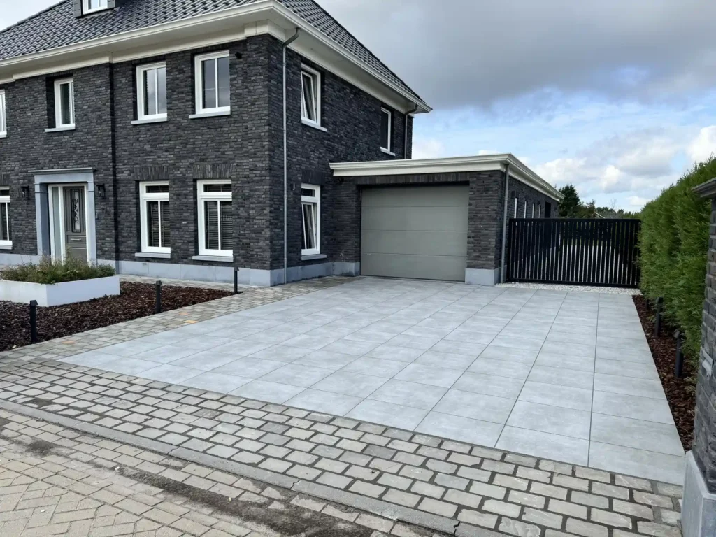 Ruime oprit met grijze terrastegels en klinkerbestrating bij een moderne woning met garage en zwart hekwerk