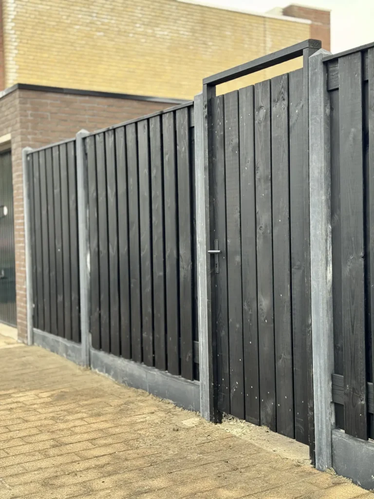 Grijze houten schutting met betonpalen langs de bakstenen gevel van een woning