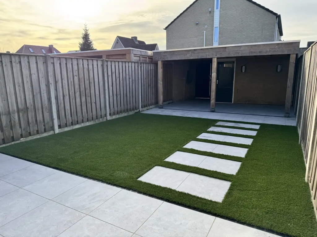 Achtertuin met kunstgras, grijze terrastegels als stapstenen, een houten overkapping en schutting bij zonsondergang