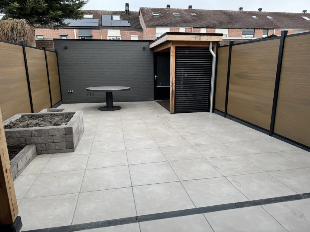 Ruim betegeld terras met grote grijze tegels, een gemetseld plantvak, overkapping en houten schuttingen