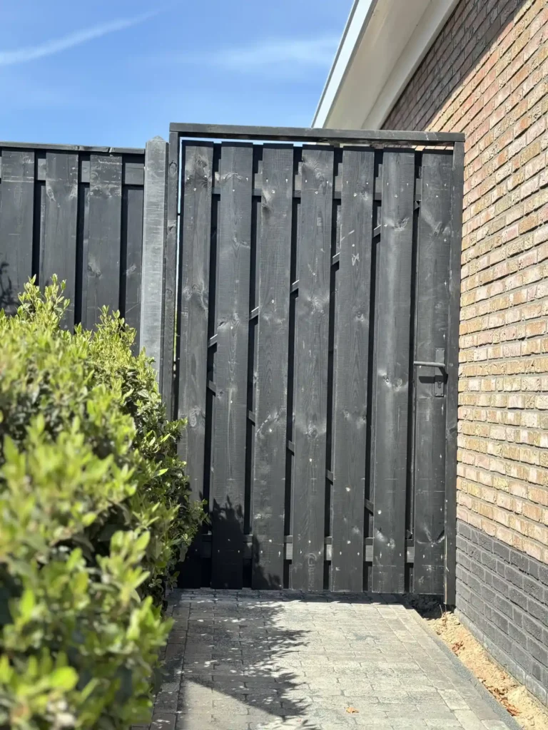Zwarte houten schutting met poort en betonpalen langs de bakstenen gevel van een woning