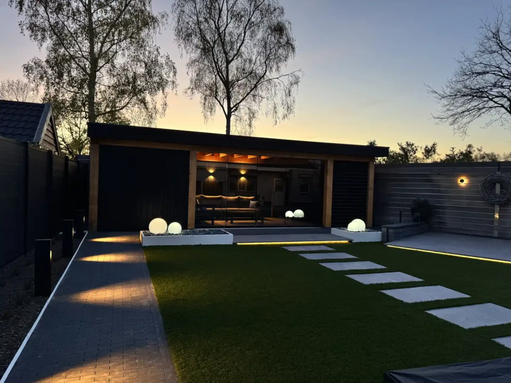 Luxe tuin bij zonsondergang met verlichte overkapping, loungeset, verlichte plantvakken en kunstgras met stapstenen