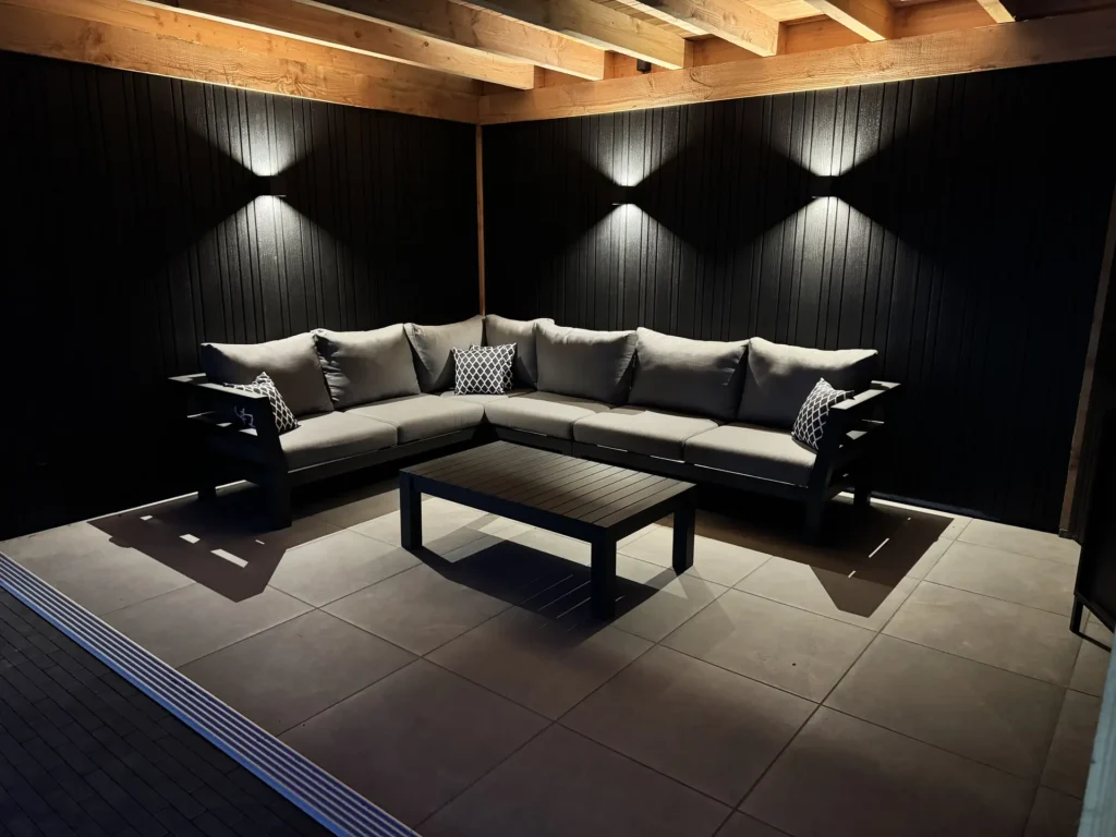 Overdekt terras met grijze loungeset, zwarte houten wanden met sfeerverlichting en een tegelvloer