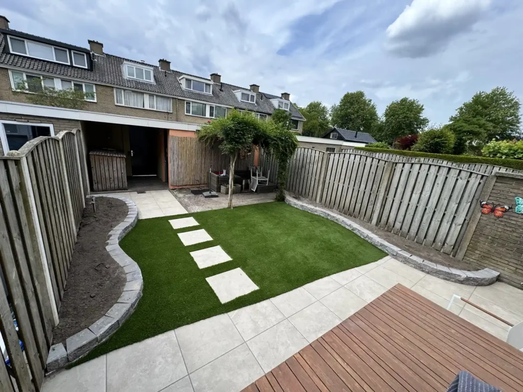 Aangelegde achtertuin met kunstgras, grijze terrastegels als stapstenen, een gebogen border en een houten vlonder
