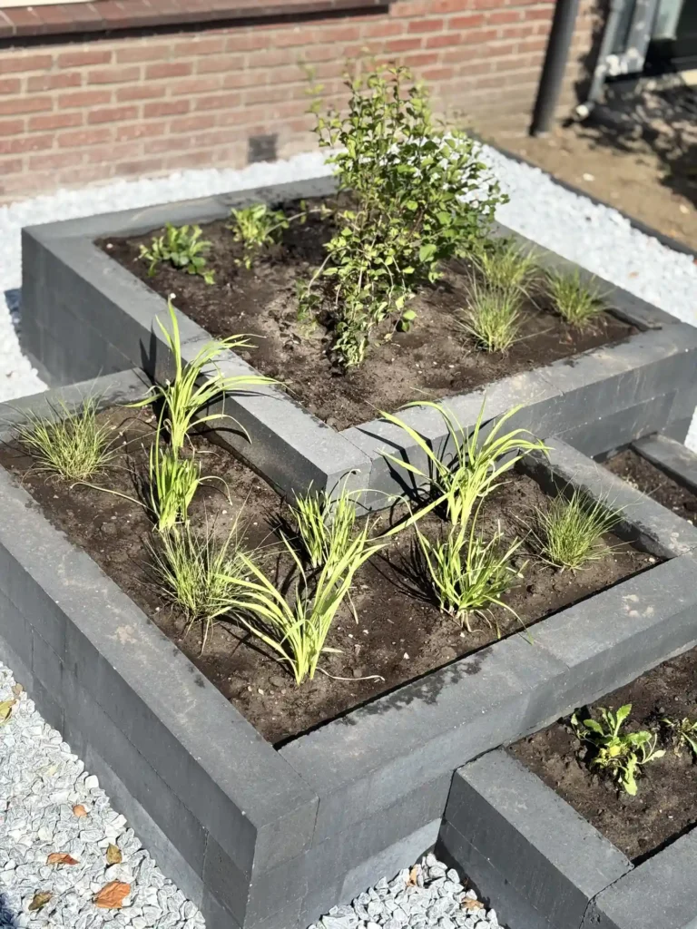 Verhoogd plantvak met antraciet betonranden, jonge beplanting en siergrind langs een bakstenen muur