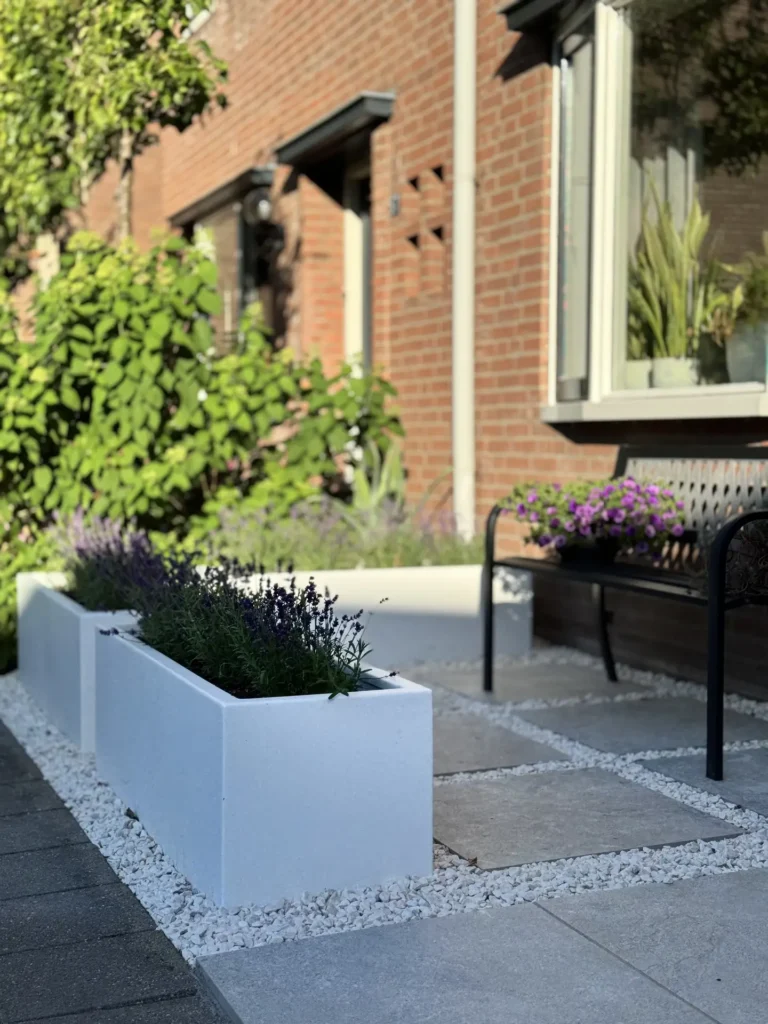 Leibomen en bloeiende beplanting langs de bakstenen gevel van een woning met tuinbank